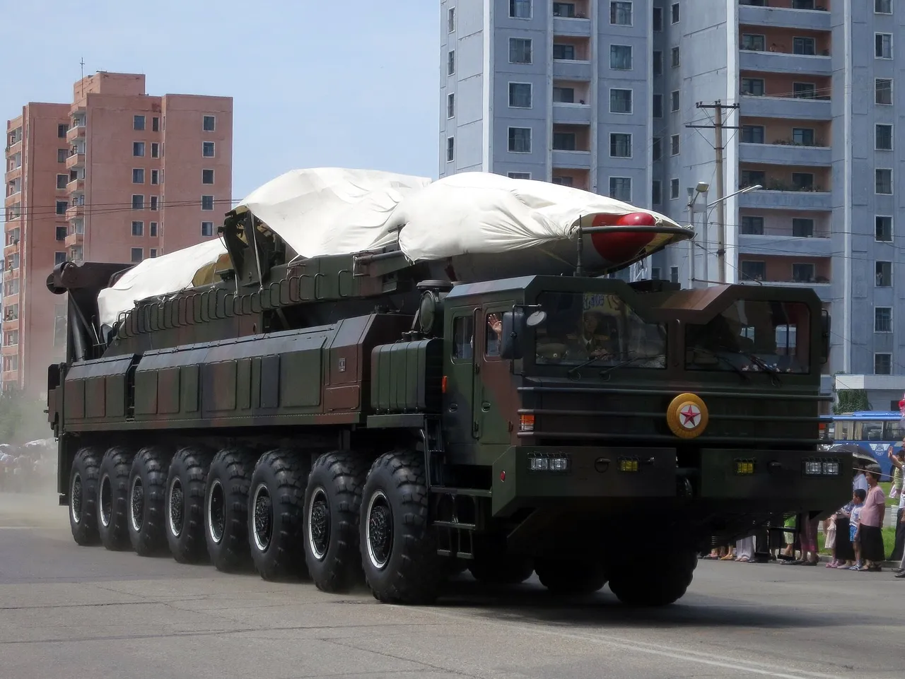 A large multi-axle green military launcher truck carrying a missile under white covers drives past apartment blocks in Pyongyang