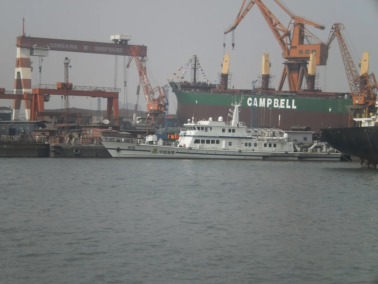 A white China Coast Guard patrol cutter, hull number 015, moored at a shipyard with cranes and a large cargo ship behind it