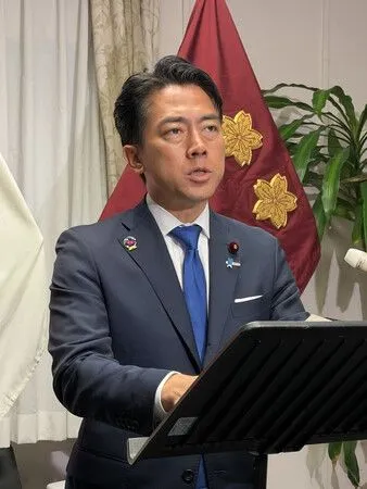 Japanese Defence Minister Shinjiro Koizumi in a dark suit and blue tie speaking at a lectern in front of a red flag