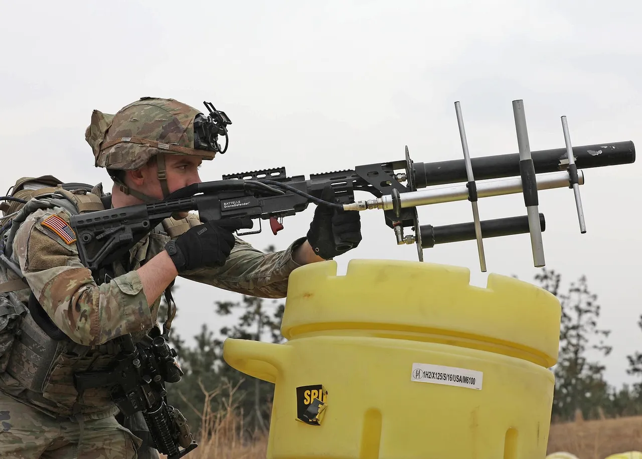 A US soldier in camouflage and helmet aims a rifle-shaped drone jammer fitted with antennas over a yellow barrier outdoors