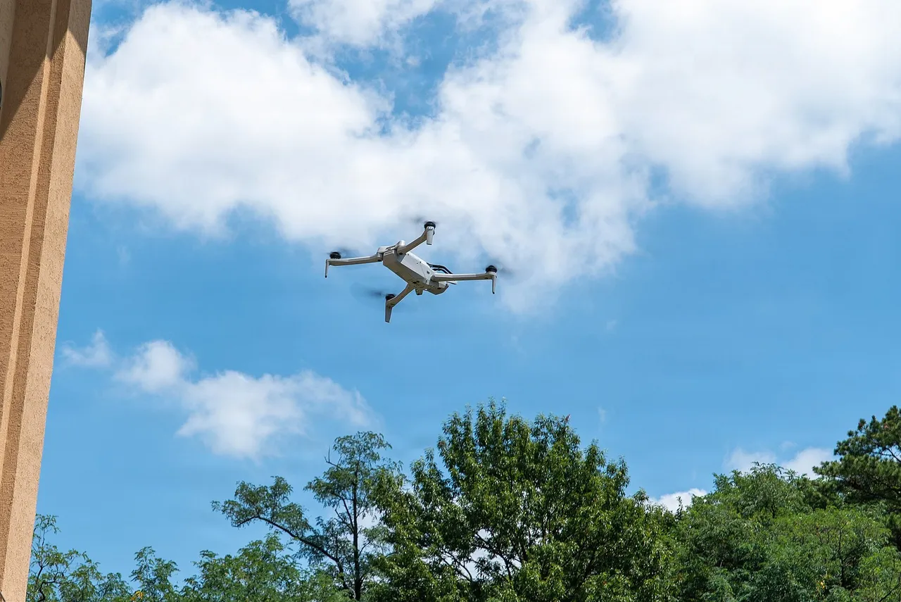 A small white quadcopter drone flies against a blue sky with white clouds above a line of green trees