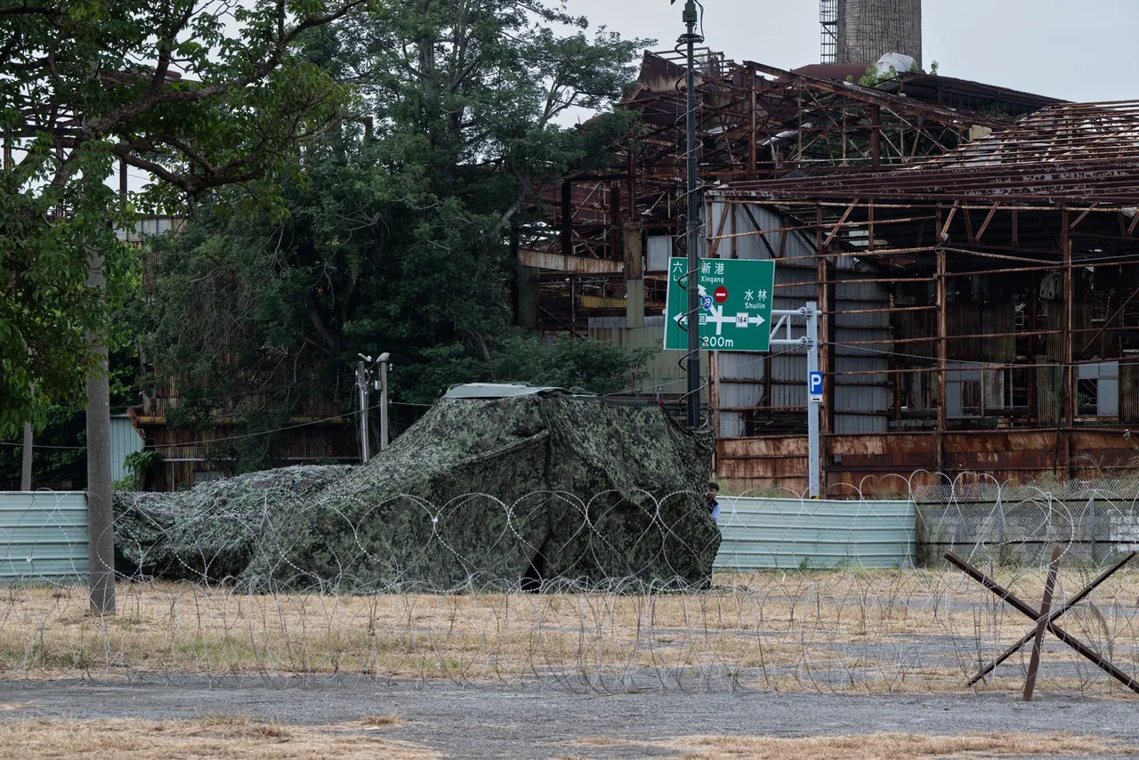 A vehicle covered in camouflage netting sits behind barbed wire and anti-vehicle obstacles in front of a ruined building.