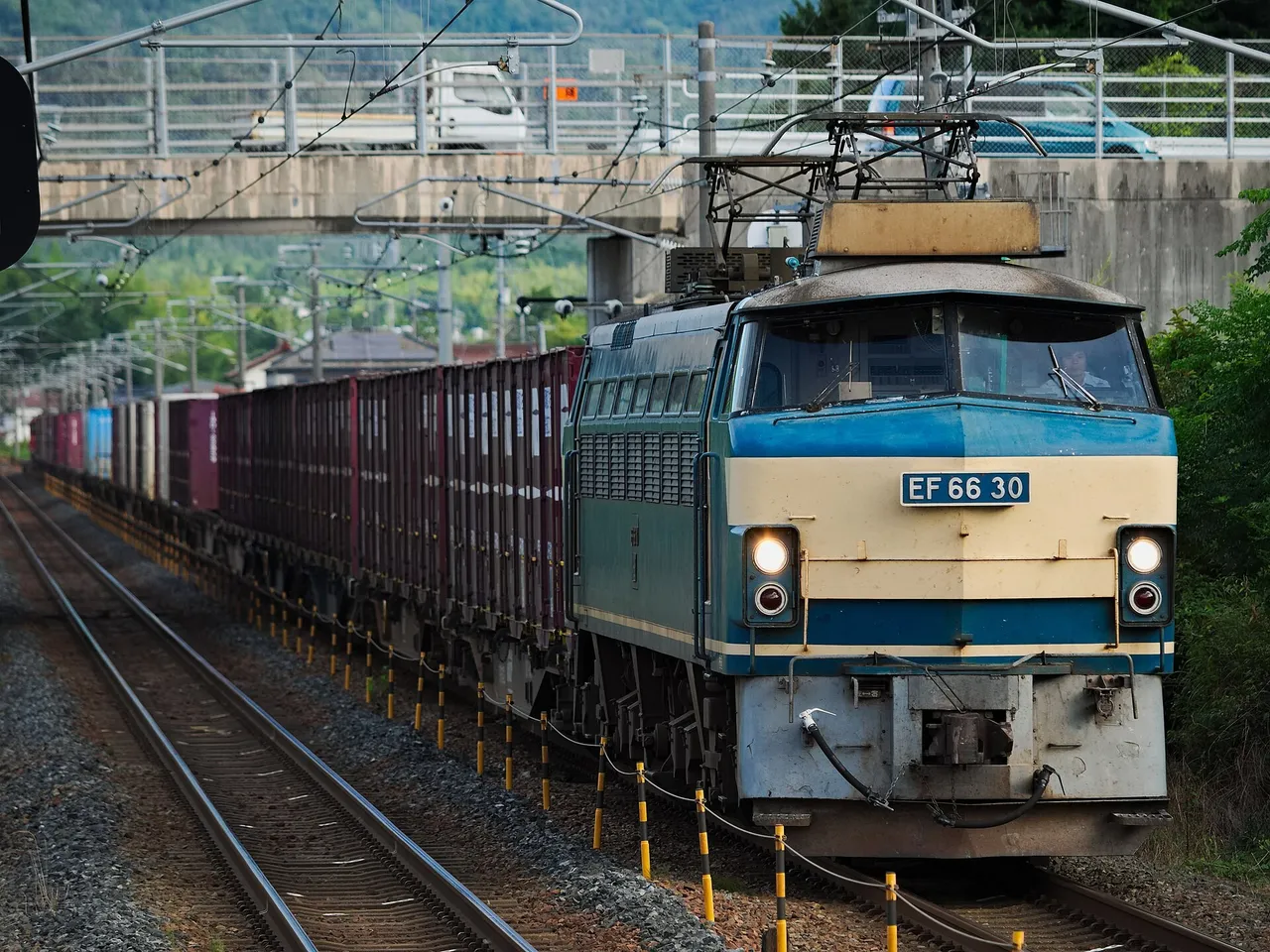 A blue and cream electric locomotive numbered EF66 30 pulls a long line of red freight containers along a double-track railway