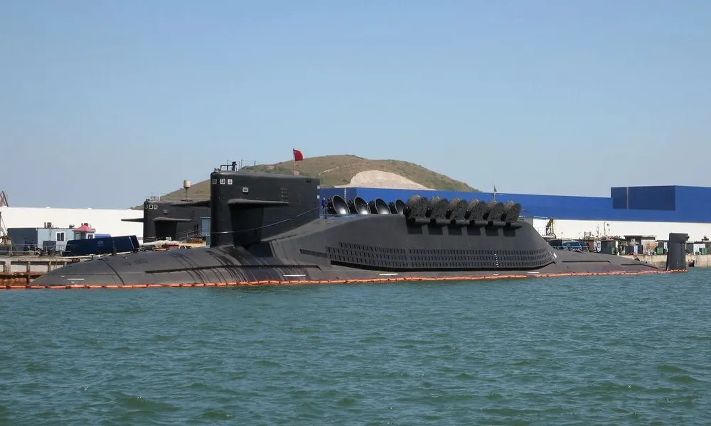 A large black submarine with a row of open missile hatches behind its sail, moored at a pier beside a blue warehouse