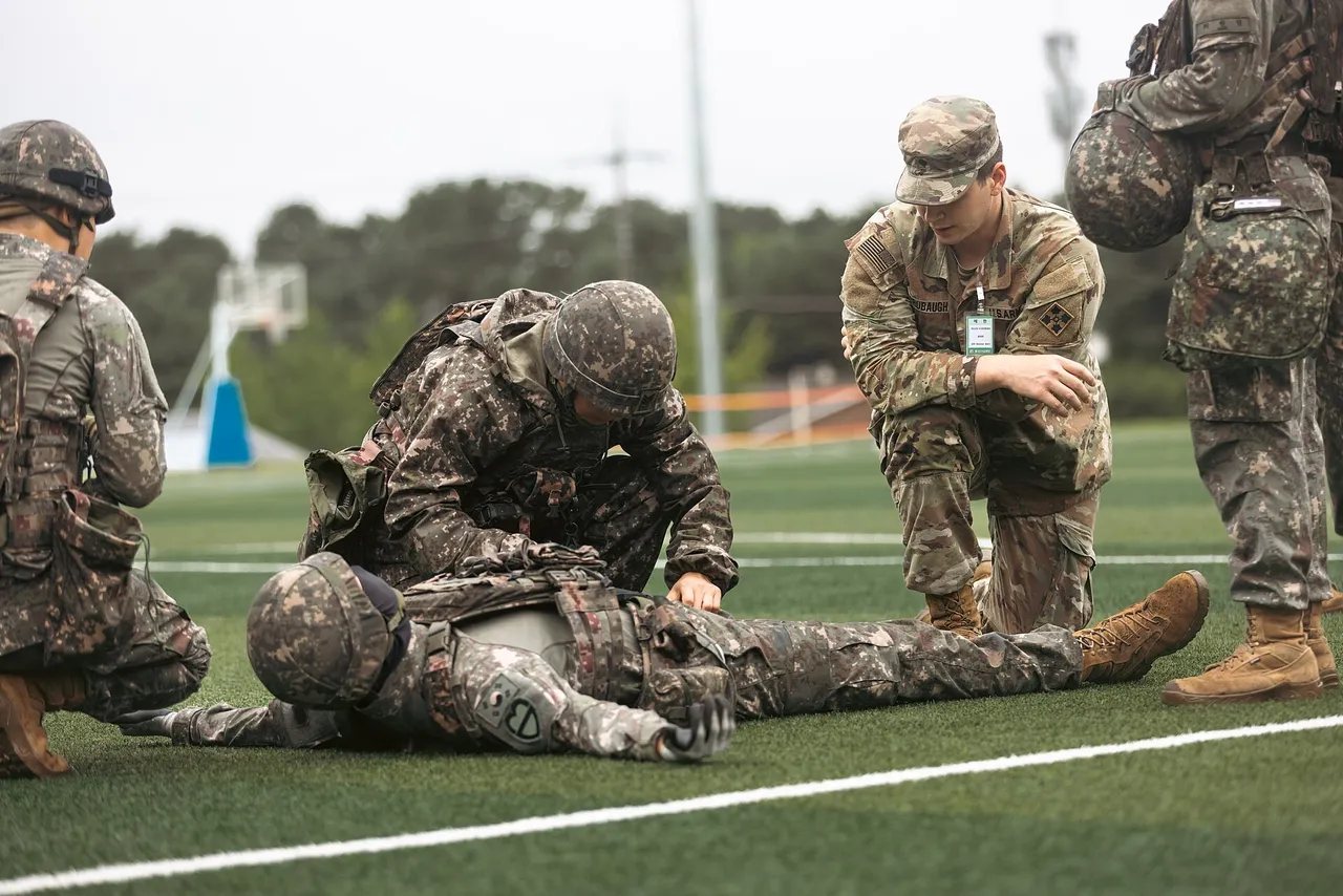 South Korean soldiers in camouflage kneel over a soldier lying on a sports field to treat a simulated wound as a US Army medic watches