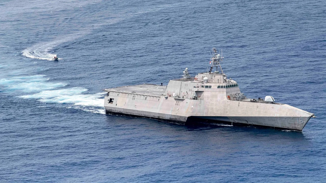 A grey angular US Navy littoral combat ship at sea with a small inflatable boat approaching in its wake