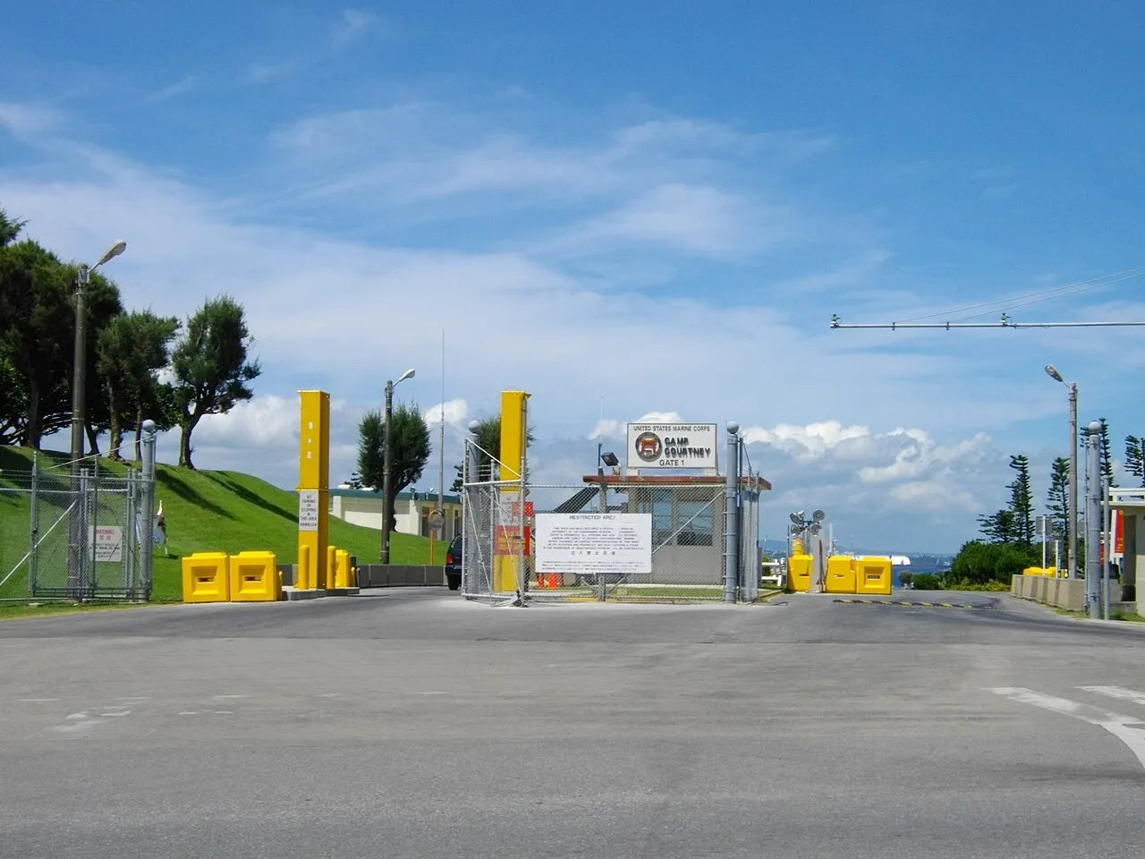 A guarded entrance gate to a US Marine Corps base with yellow barriers, a sign reading Camp Courtney Gate 1, a fence and a small guardhouse under a blue sky.
