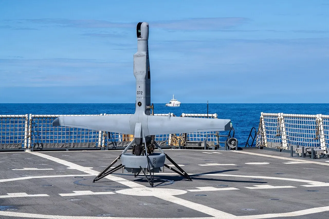 A grey tail-sitting drone with straight wings stands upright on four legs on a ship's flight deck, with blue ocean and a distant white vessel behind.