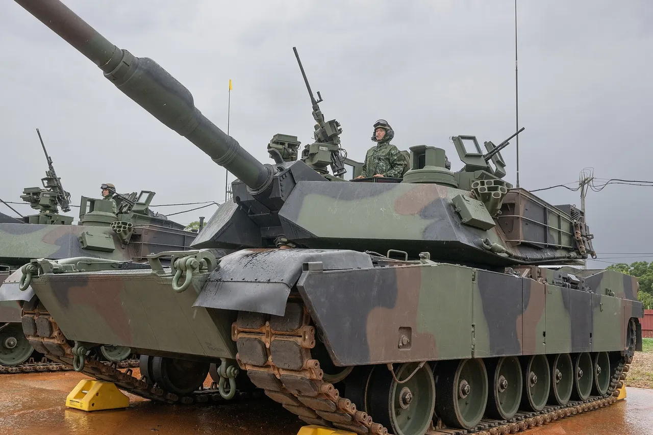 A camouflaged M1A2T Abrams tank with its long gun barrel raised and a soldier in the turret hatch, with a second tank behind under a grey sky