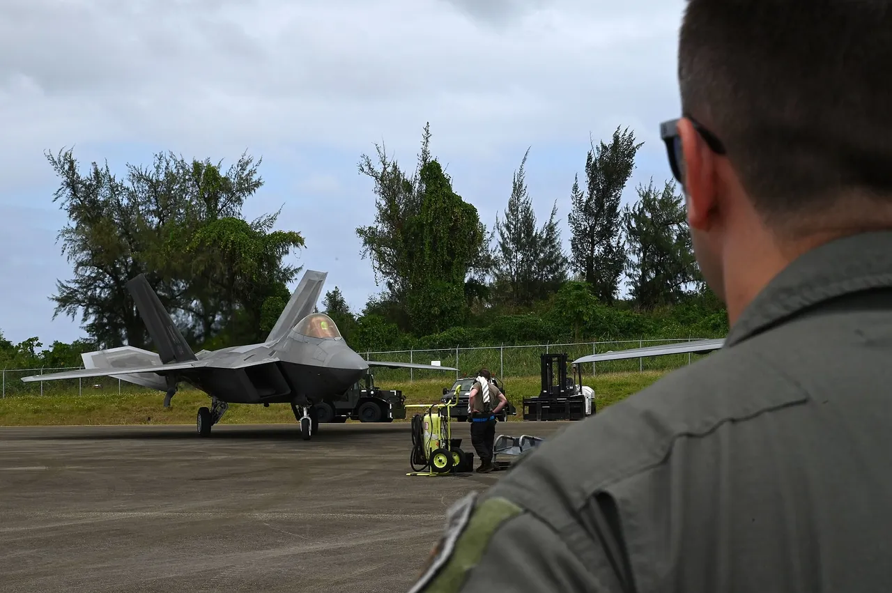 An F-22 stealth fighter taxis on an island airfield lined with trees as a US airman in a flight suit watches in the foreground