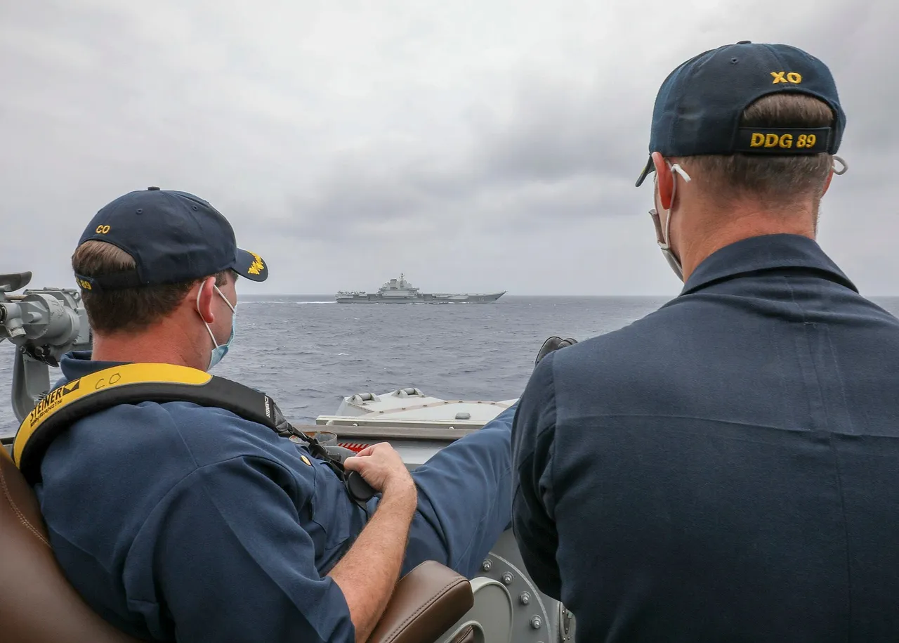 Two US Navy officers in caps seen from behind on a destroyer bridge wing, looking out at a grey aircraft carrier on the horizon