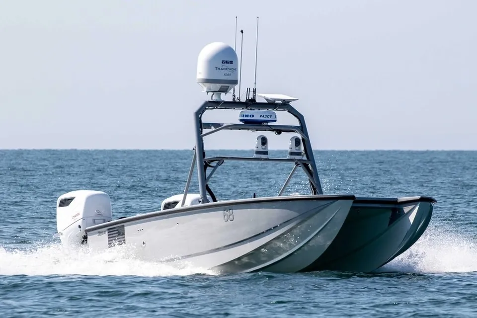 A small grey uncrewed speedboat with a sensor mast cuts across open water, leaving a white wake