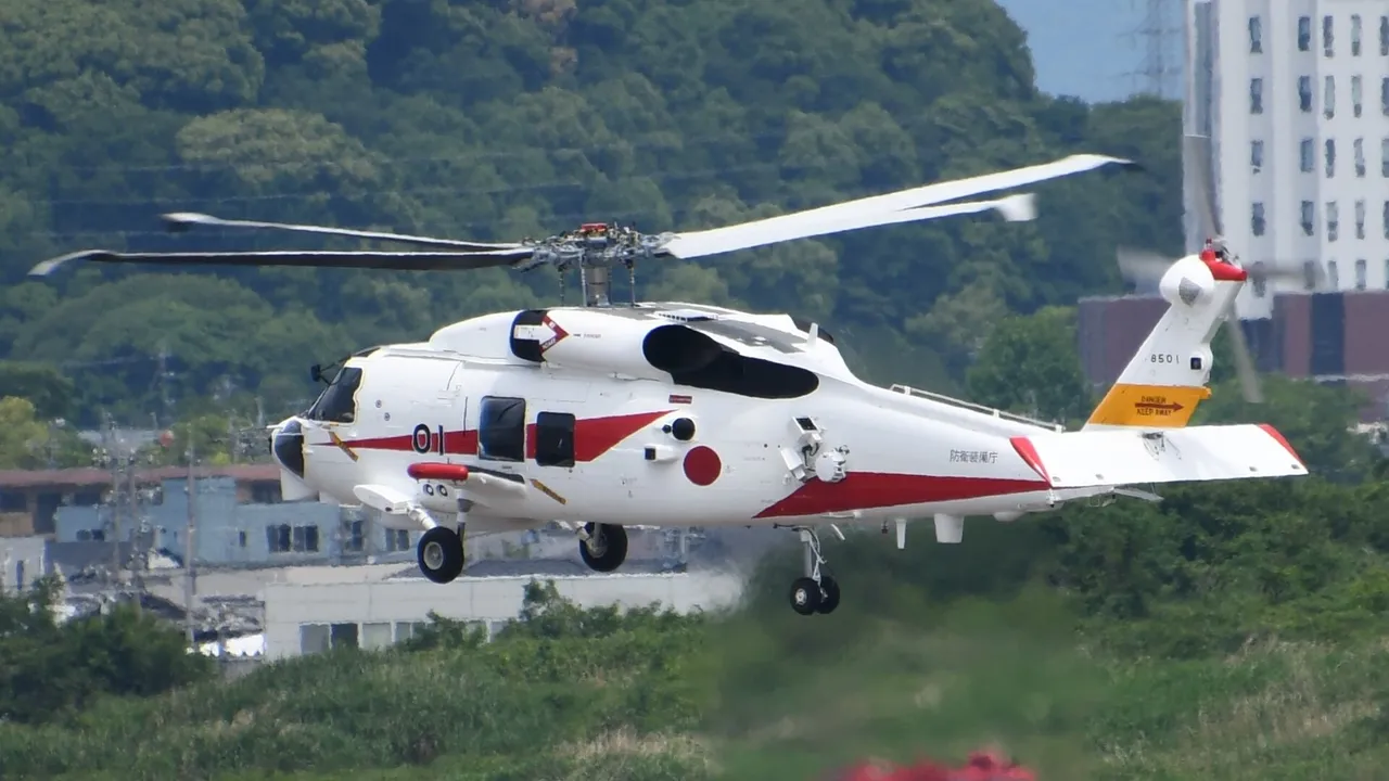 A white helicopter with red stripes and a red roundel hovers low over grass in front of factory buildings and wooded hills