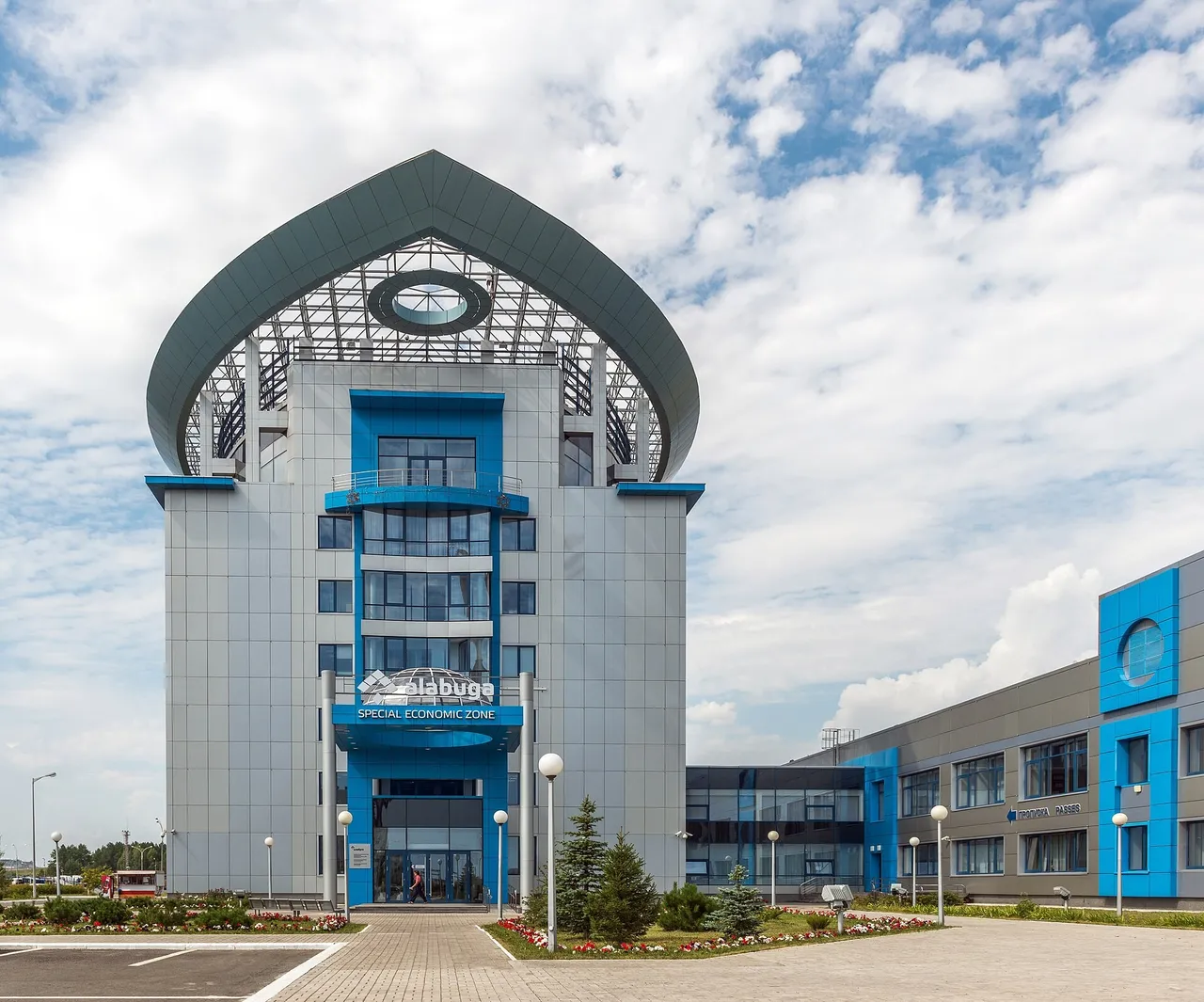 A tall grey and blue office building with an arched glass roof and an Alabuga Special Economic Zone sign at its entrance