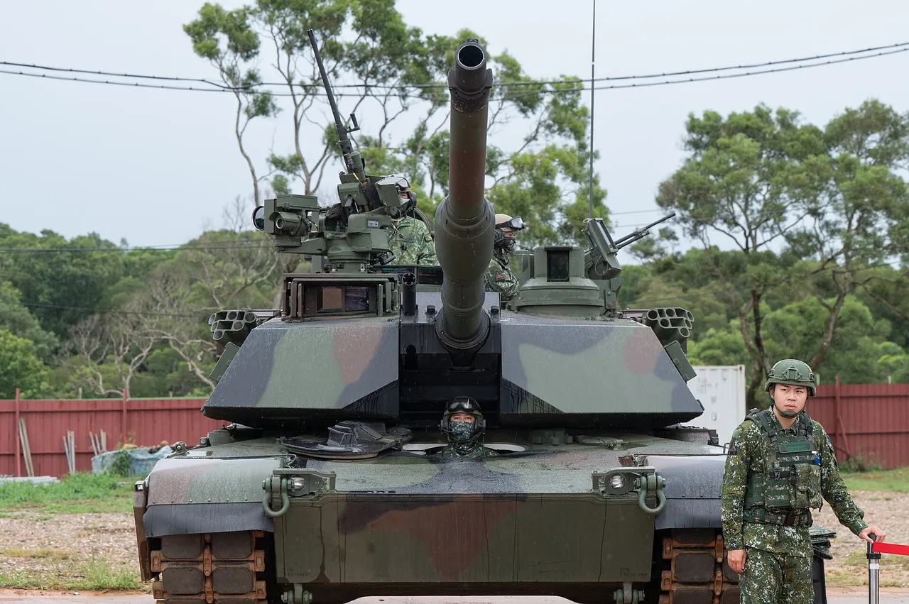 A camouflaged M1A2T Abrams tank with soldiers in the hatches and a soldier standing beside it