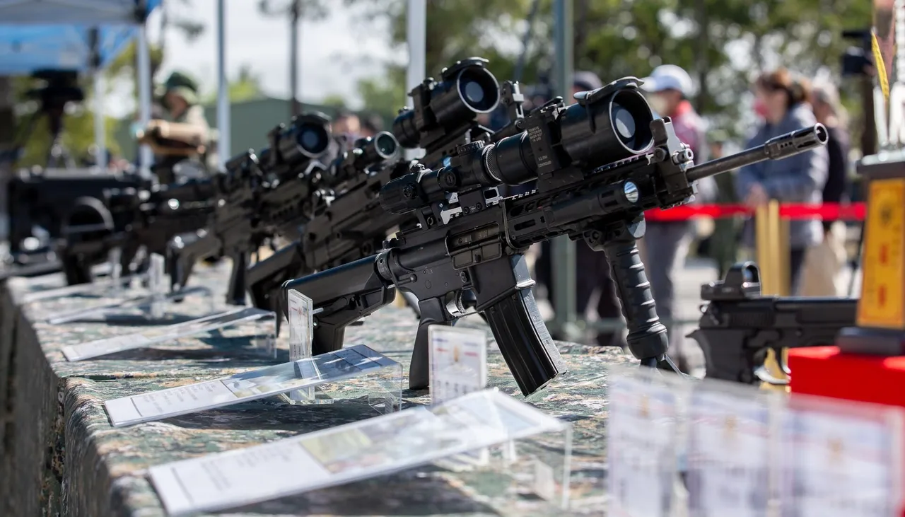 A row of black assault rifles fitted with optical sights displayed on a camouflage-covered table outdoors