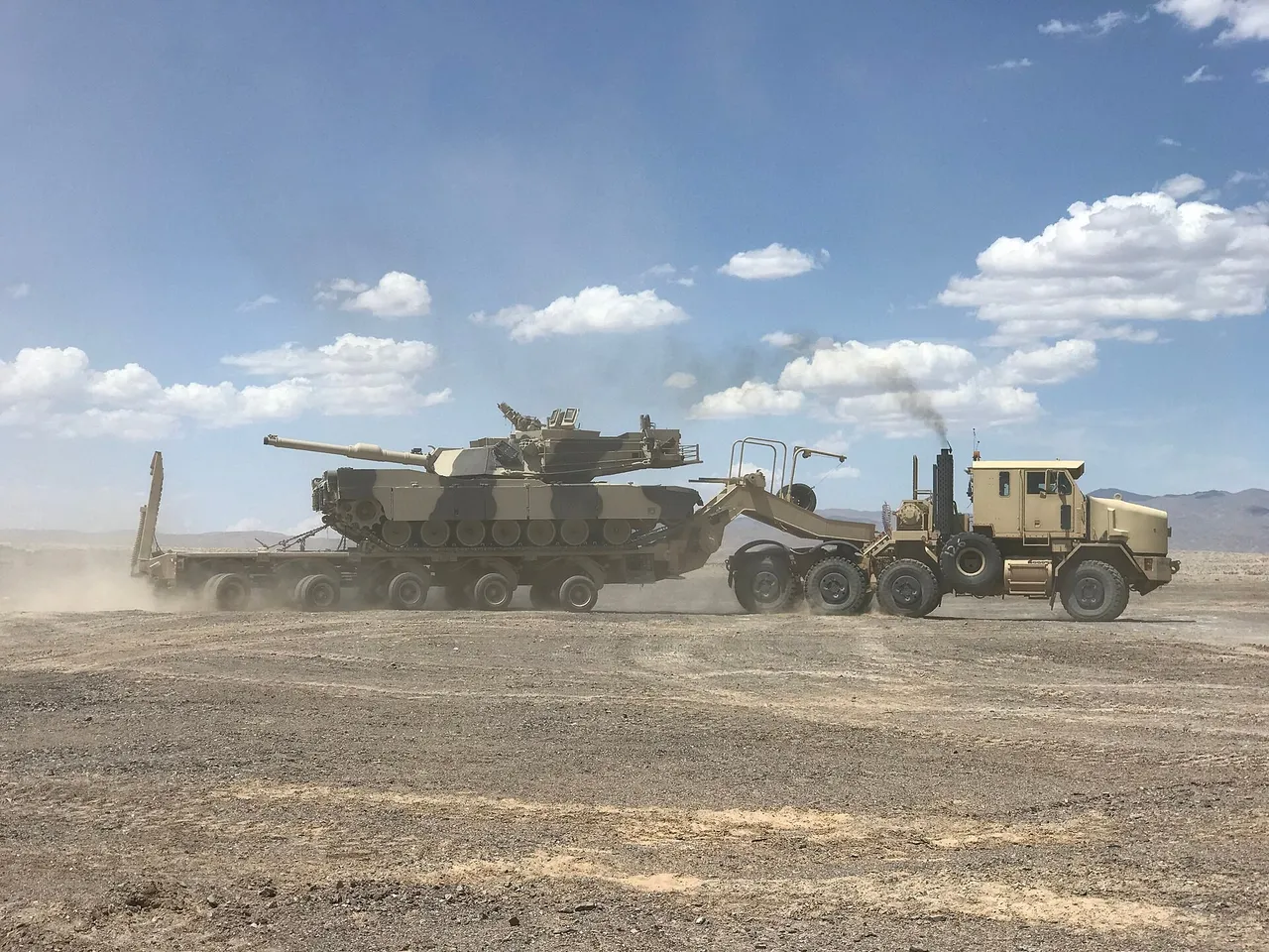 A desert-tan M1070A1 truck tows a long flatbed trailer carrying an M1 Abrams tank across dusty desert ground