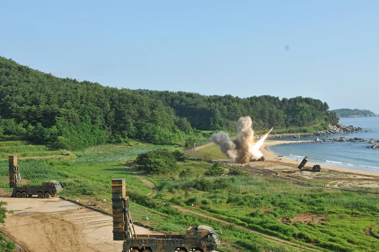 Two South Korean missile launcher trucks with raised canisters on a green coastal hillside as a rocket launches in a cloud of smoke near the beach