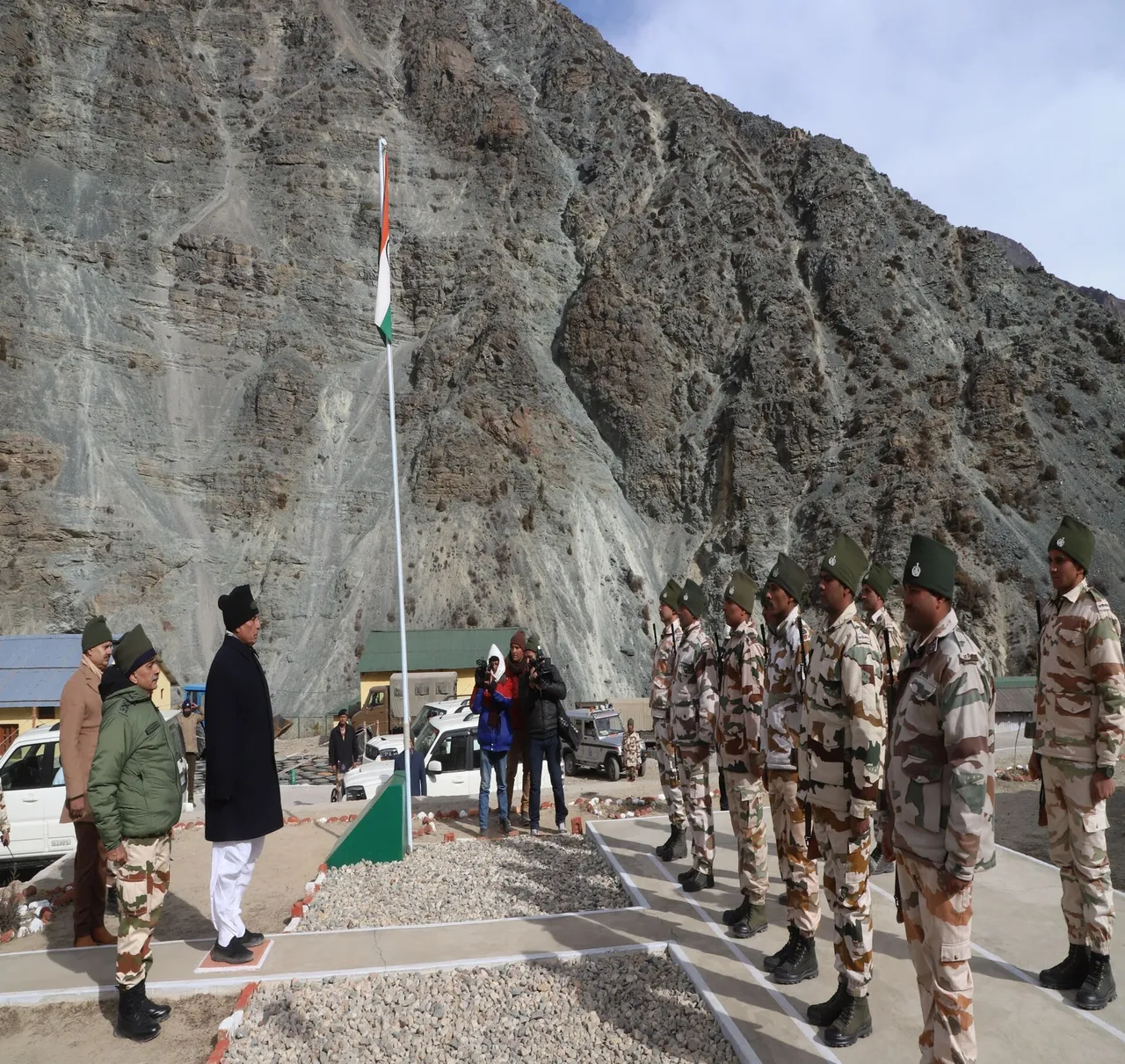 Indian border police in camouflage uniforms and green caps stand in a line beside a flagpole at a mountain outpost below a steep rocky slope