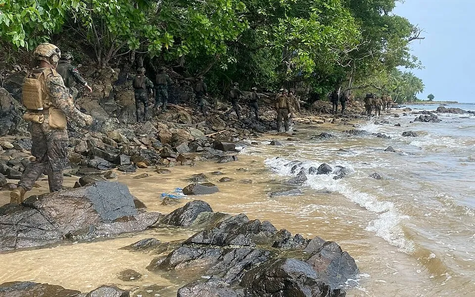 Marines in camouflage and helmets walk in a line along a rocky, tree-lined beach as small waves wash ashore.