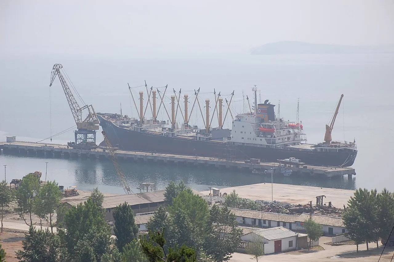 A large cargo ship moored at a long concrete pier with cranes, seen through haze from a hillside above low warehouse buildings and trees