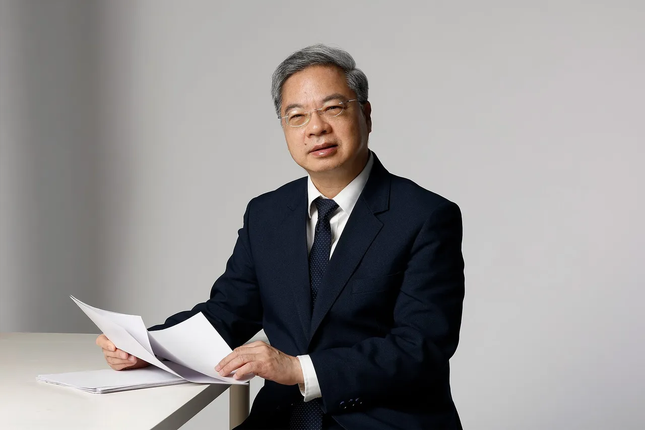 Kung Ming-hsin, in a dark suit and glasses, sits at a white desk holding papers against a grey background.
