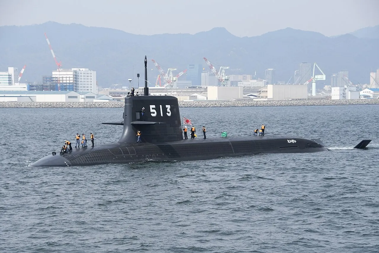 JS Taigei, a grey diesel-electric attack submarine with the sail number 513, sails outbound past industrial waterfront buildings.