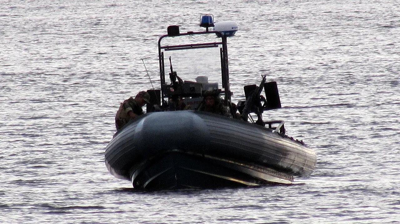 A dark grey rigid-hull inflatable boat with armed crew members aboard moves across calm grey water