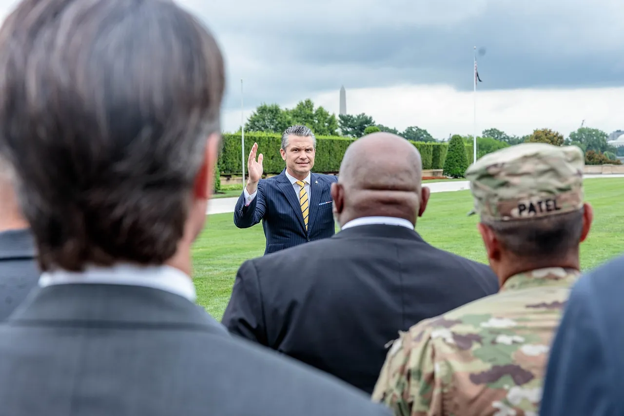 Pete Hegseth in a suit raises his hand while speaking to service members and officials on a Pentagon lawn