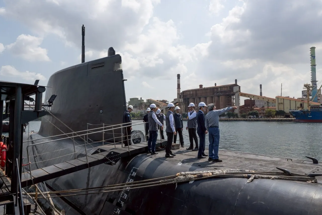 A group of people in white hard hats stand on the black deck of a submarine beside its tall sail, with a harbour and industrial buildings behind them