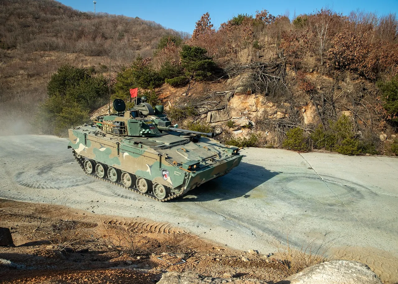 A camouflaged tracked armoured vehicle with a turret and a small red flag drives on a dusty concrete track below a rocky, wooded hillside