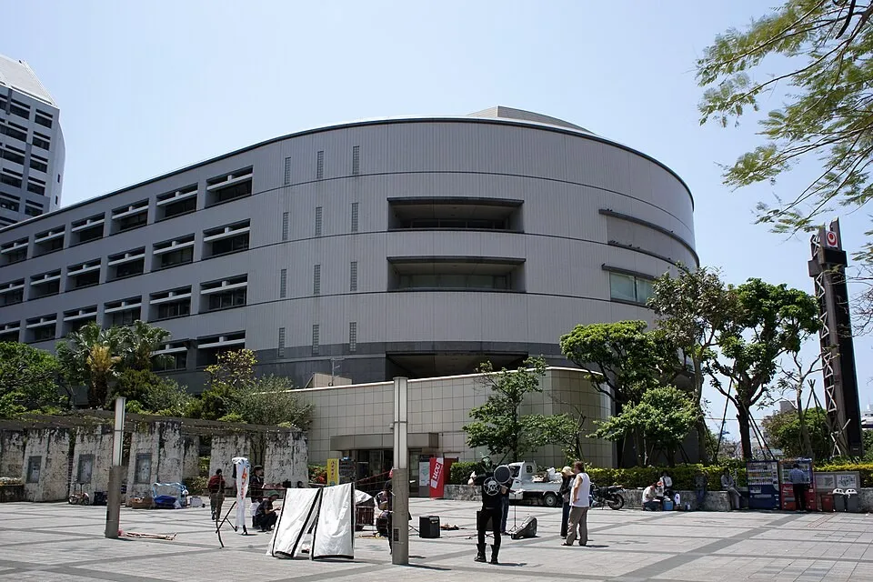 Curved grey multi-storey Okinawa Prefectural Assembly building under a clear sky, with trees and people on a paved plaza in front